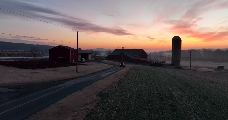 Truck drives on rural road past farm buildings and barn in morning sunrise. Turn signal. Aerial.