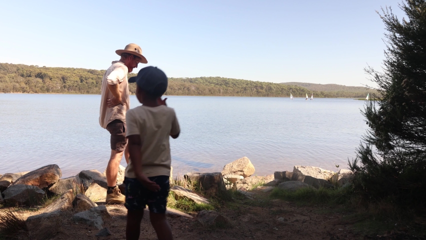A man with a hat and ginger beard and his son look out at a lake with boats on it.