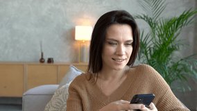 Happy pleasant woman relaxing on comfortable couch, holding smartphone in hands. Smiling young lady chatting in social networks, watching funny videos, using mobile applications at home.