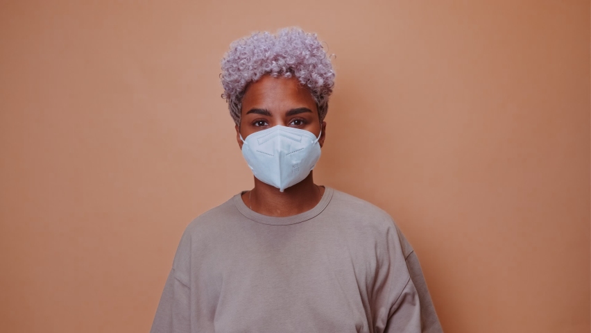 Young puzzled ethnic African American woman in blue gloves with respirator on face, clutching head from what he saw, preparing to clean foul-smelling room stands on brown background