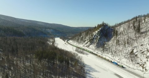 Freight long train carries with cargo carriages in hard to reach wild mountains landscape through a difficult part of Trans Siberian railways. Aerial drone view at winter sunny day.