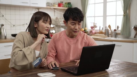 happy asian married couple enjoying shopping online with credit card and laptop. the man is keying in the number to make payment as the woman is reading to him