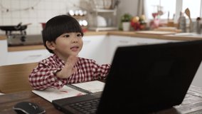 cute Asian schoolboy waving hi to laptop screen while taking online lesson in the dining room at home. learning from home concept