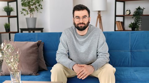 Confident handsome hipster bearded man in eyeglasses sitting on blue couch at cosiness living room interior. Successful trendy male positive emotion wearing fashion gray sweatshirt relaxing at home 