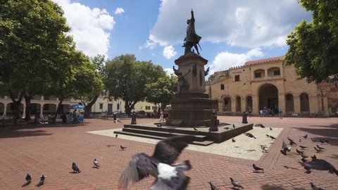 Colon Park, Santo Domingo, Dominican Republic - March 15, 2022. Stone-paved ancient square of a big city. City landscape of sights. Birds on the monument. Green trees in the park.