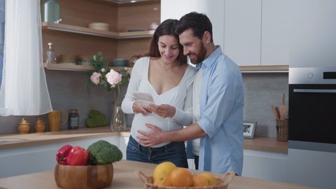 Loving caucasian young family husband and pregnant wife looking at ultrasonography child x-ray photograph on the kitchen. Happy aspiring parents couple at cozy house.