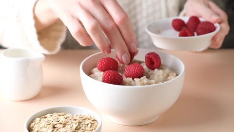 Oatmeal porridge with raspberries for breakfast. Woman adding fresh raspberries into cooked oatmeal bowl