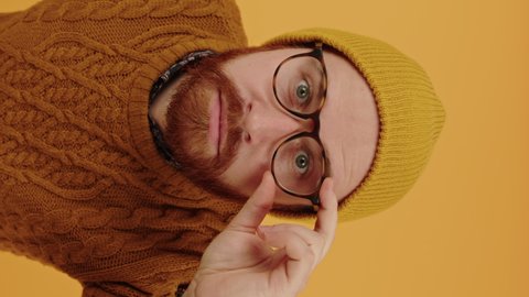 Young suspicious Caucasian man bends down to the camera, adjusts glasses, shakes head no sign trying to see something closeup vertical video isolated yellow background . High quality 4k footage