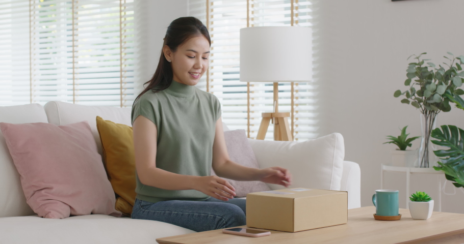 Young happy asia people teen girl smile enjoy unbox post mail sit relax at home comfort sofa couch feel joy amazed awe wow in omni channel fast send parcel via online sale shop store e-commerce order. | Shutterstock HD Video #1088493155