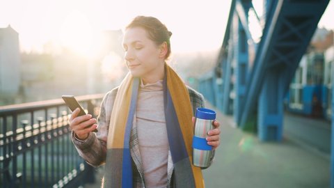 Portrait of a young caucasian businesswoman in a coat, walking across the bridge on a frosty morning, drinking coffee and using smartphone. Communication, work day, busy life concept.