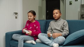 Moving shot of diverse kids playing video game while sitting on sofa indoors. Multiethnic preteen friends having fun at home, spending leisure time together. Children relaxing at home