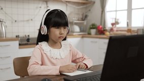 Taiwanese girl with headphones waving hi at computer screen and having conversation with teacher through video for a language learning lesson at dining table. e-learning at home concept