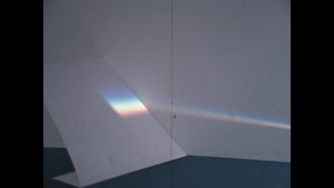 1960s: Shaft of light shines across background. Man moves prism into path of light. Light shines a rainbow on piece of paper across table.