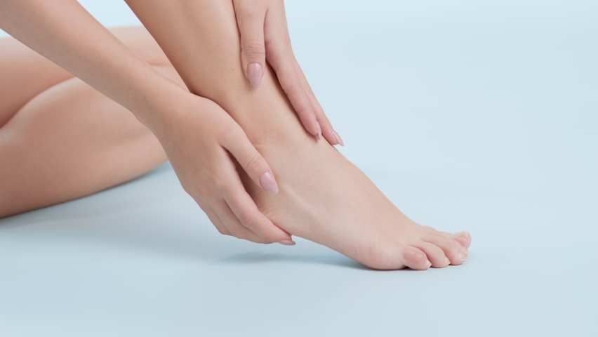 Horizontal close-up shot of gorgeous young slim woman in black top strokes her foot and leg sitting on the floor on pale blue background Foot and leg care concept Royalty-Free Stock Footage #1088511093