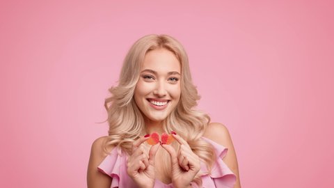 Smiling Blonde Lady Holding Two Heart Shaped Jelly Candies Posing Over Pink Studio Background. Love And Romantic Relationship. Sweet Tooth Concept. Slow Motion