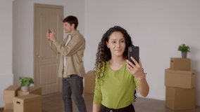 New House. Cheerful Spouses Video Calling Via Smartphones Celebrating Moving In Together Standing Among Cardboard Boxes At Home After Relocation. Real Estate Purchase And Ownership Concept