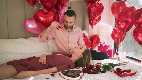 Romantic couple petting in bed. Young attractive woman lying with closed eyes, man gently throwing red roses petals on her body. Marriage, family, love, sex, intim concept. RED camera shot slow motion