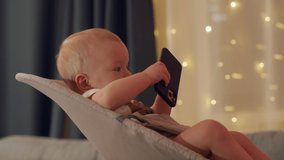 Baby boy sitting on rocking chair for babies playing with smartphone, cute toddler watching cartoon on mobile phone screen, child and modern technology concept. High quality 4k footage