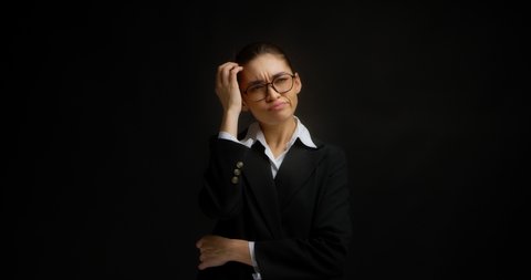 A confused businesswoman in glasses and office clothes scratches her head, holds her chin with her hand, remembers something and thinks about it. Isolated on a black background