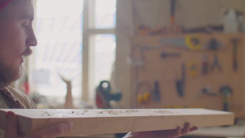 Woodworker blowing dust from wooden plank and examining it while working in carpentry workshop