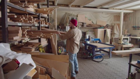 Artisan taking plank from shelf, then bringing it to workbench and putting on safety earmuffs before woodworking in carpentry workshop