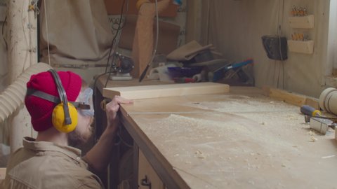 Woodworker in safety earmuffs and glasses shaping wooden plank on trimmer table in carpentry workshop