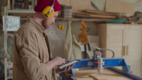 Woodworker in safety earmuffs using smartphone in workshop while operating woodworking CNC machine with mobile app