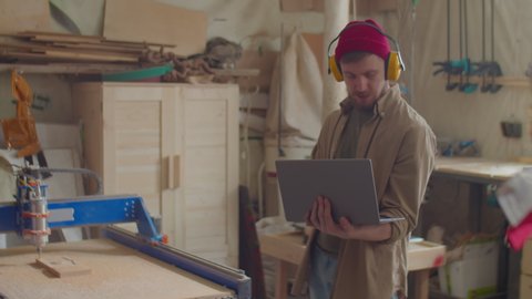 Woodworker in safety earmuffs standing in workshop and operating CNC machine with laptop