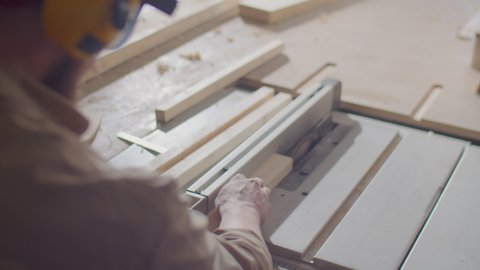 Professional woodworker in safety earmuffs and glasses cutting wooden board with table saw while working in carpentry workshop