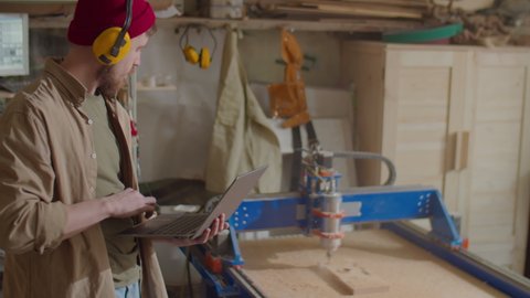Carpenter in protective earmuffs using laptop while operating woodworking CNC machine in workshop