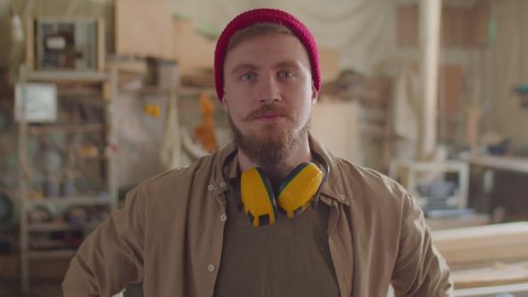 Portrait of bearded woodworker with safety earmuffs over his neck posing for camera in carpentry workshop