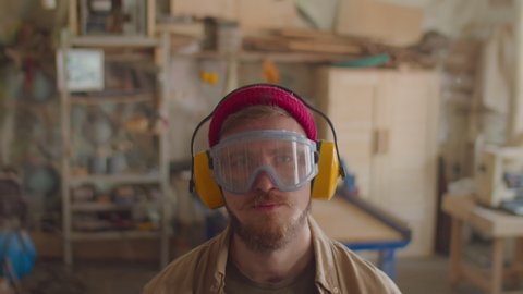 Portrait of Caucasian male carpenter in safety earmuffs and glasses posing for camera in woodworking workshop