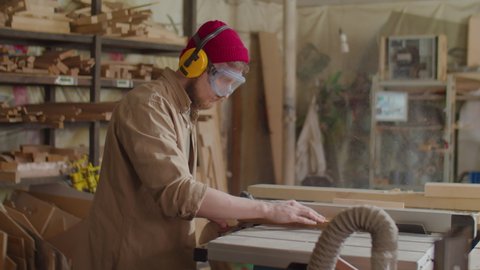 Male carpenter in safety earmuffs and glasses using table saw for cutting plank in woodworking workshop