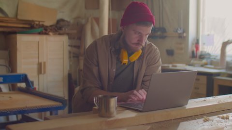 Young bearded woodworker with safety earmuffs over his neck browsing the Internet on laptop at workbench in carpentry workshop