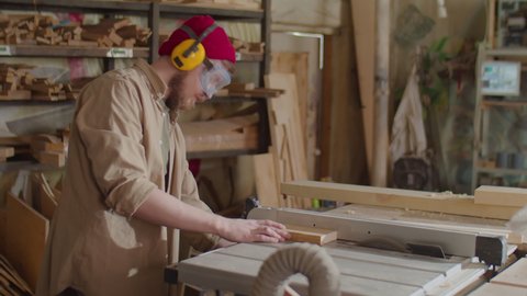 Carpenter in safety earmuffs and glasses cutting board with table saw in woodworking workshop