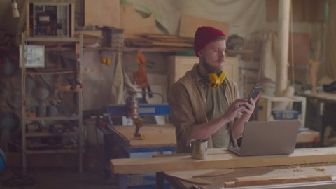Young male woodworker with safety earmuffs over his neck using smartphone in carpentry workshop