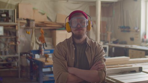 Portrait of woodworker in safety earmuffs and glasses posing for camera with arms crossed in carpentry workshop