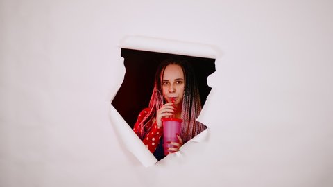 Barmaid with drink looking through hole in paper background. Female bartender with dreadlocks and cup of sweet beverage peeking through ripped white paper background in studio and looking at camera