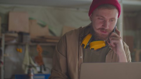 Professional woodworker with safety earmuffs over his neck talking on mobile phone and typing on laptop in carpentry workshop