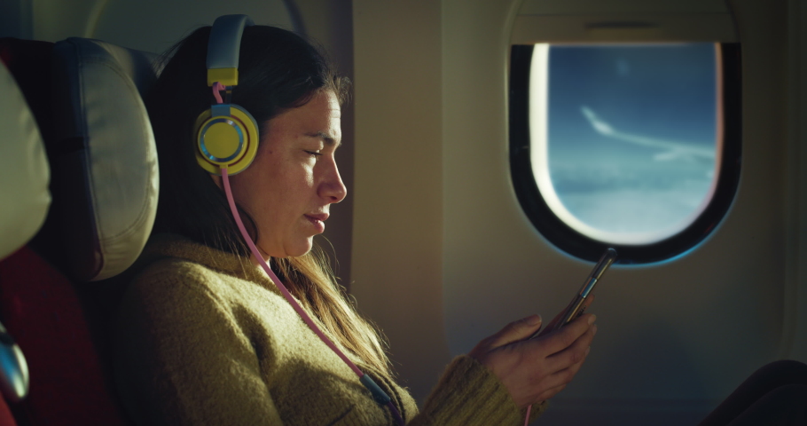 Cinematic shot of young woman with headphones listens favorite music with smartphone by using wireless connection in aircraft while traveling with comfort during international flight to vacation trip. Royalty-Free Stock Footage #1088531735