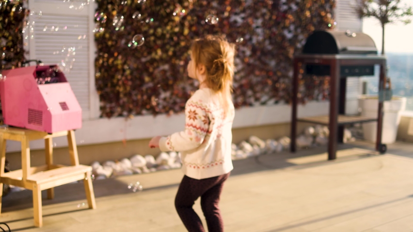 excited joyful baby girl catching and popping the flying bubbles from bubble machine, having fun on courtyard, home entertainment