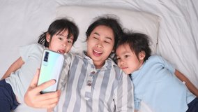 Happy Asian family enjoying with smartphone at cozy home. Smiling mother and cute daughters using phone, Watch video games or cartoons online in bed at home. Good time at home