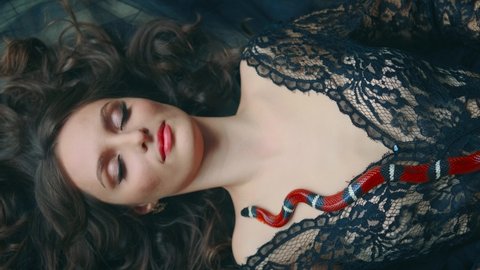 Gothic fantasy woman dark queen of snakes and poisons. Girl vampire sleeping. Beauty princess lies, hugs red snake sinaloa. Flowing hair, black dress. Royal milk snake crawls on chest. 4k footage.