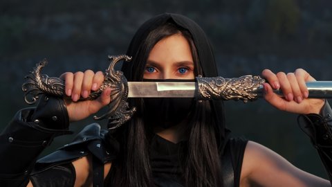 close-up portrait fantasy woman warrior assassin holding dagger in hands, hiding face behind mask. Armed girl, blue eyes. Black leather costume hood on head. Ninja soldier with knife sword. 4k footage