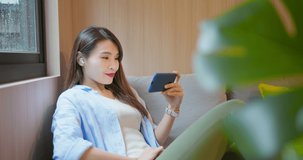 asian pretty woman lying on couch use wireless earbuds to watch comedy or video and movie by window happily at home