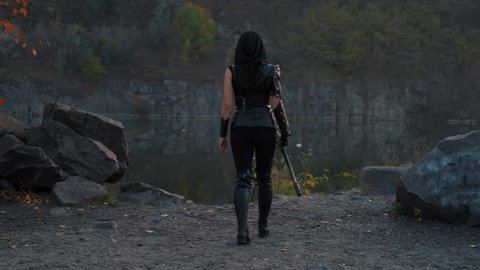 silhouette fantasy woman warrior assassin holds dagger in hands walks at night nature. Rear view, back armed girl. Black leather costume, hood. Ninja soldier with sword knife. 4k footage. river rocks