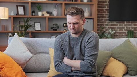 Man sitting on the couch at home, suddenly feeling strong stomach ache, gastritis problem Caucasian man squeezing belly with hands because of abdominal pain. Adult guy suffering from stomach ache.