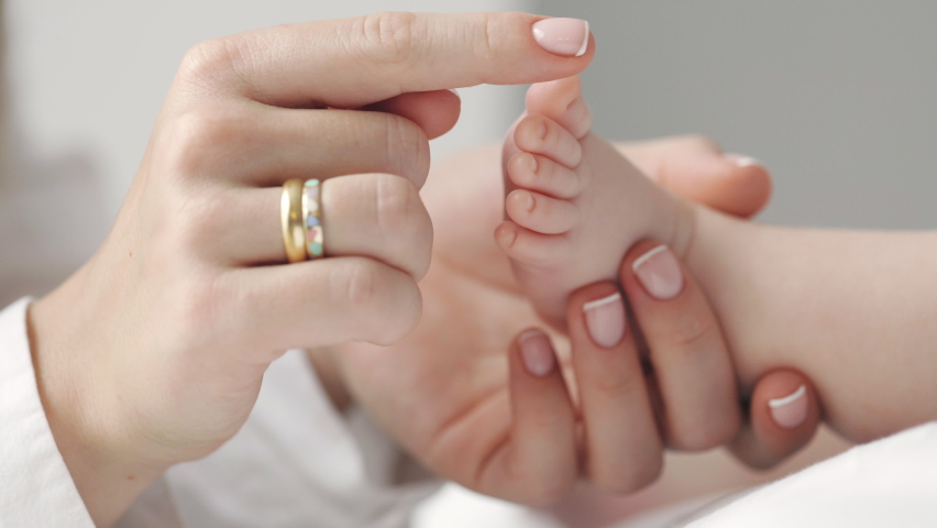 Caucasian married woman holding bare feet of her baby son and touching little fingers one by one. Caring mother enjoying time with her newborn child. Parenthood, childhood, life, maternity