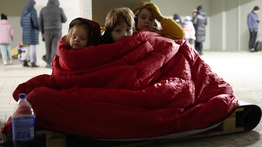 group of little kids, siblings sitting quietly on wooden pallet on the floor in bomb proof shelter during air-raid attack. Sharing one blanket during freezing night. russian invasion to Ukraine