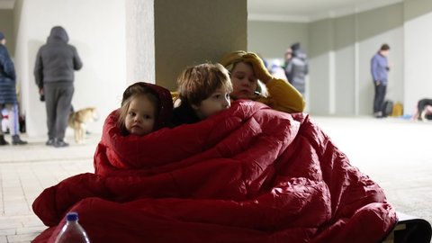 group of little kids, siblings sitting quietly on wooden pallet on the floor in bomb proof shelter during air-raid attack. Sharing one blanket during freezing night. russian invasion to Ukraine